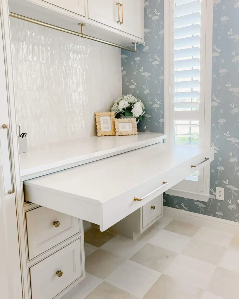 Elegant pull-out laundry countertop folding station with built-in white cabinetry, brass hardware, tiled backsplash, and soft blue wallpaper, offering a space-saving and functional laundry room design.