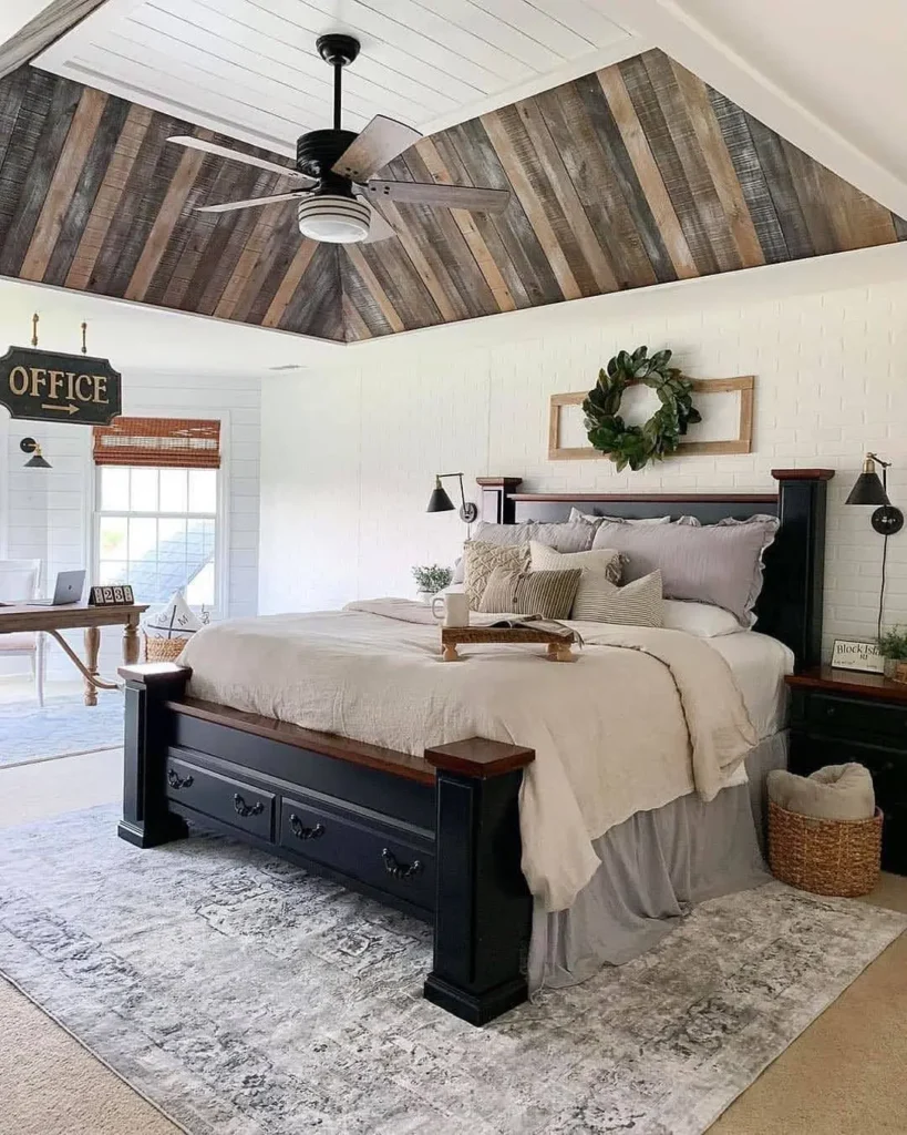 Rustic farmhouse bedroom with a black wooden bed frame, neutral layered bedding, reclaimed wood vaulted ceiling, ceiling fan, soft area rug, and cozy farmhouse accents.