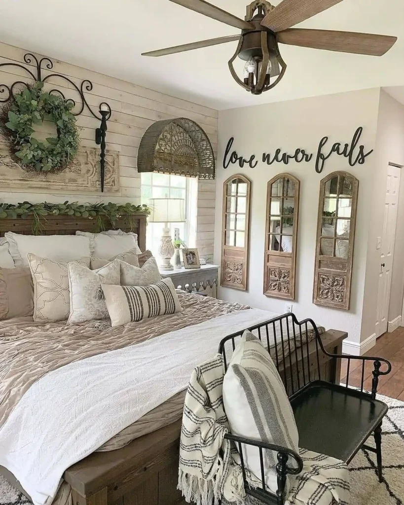 Rustic industrial farmhouse bedroom with a wood bed frame, layered neutral bedding, shiplap accent wall, vintage wall decor, metal bench, ceiling fan, and cozy farmhouse bedroom styling.