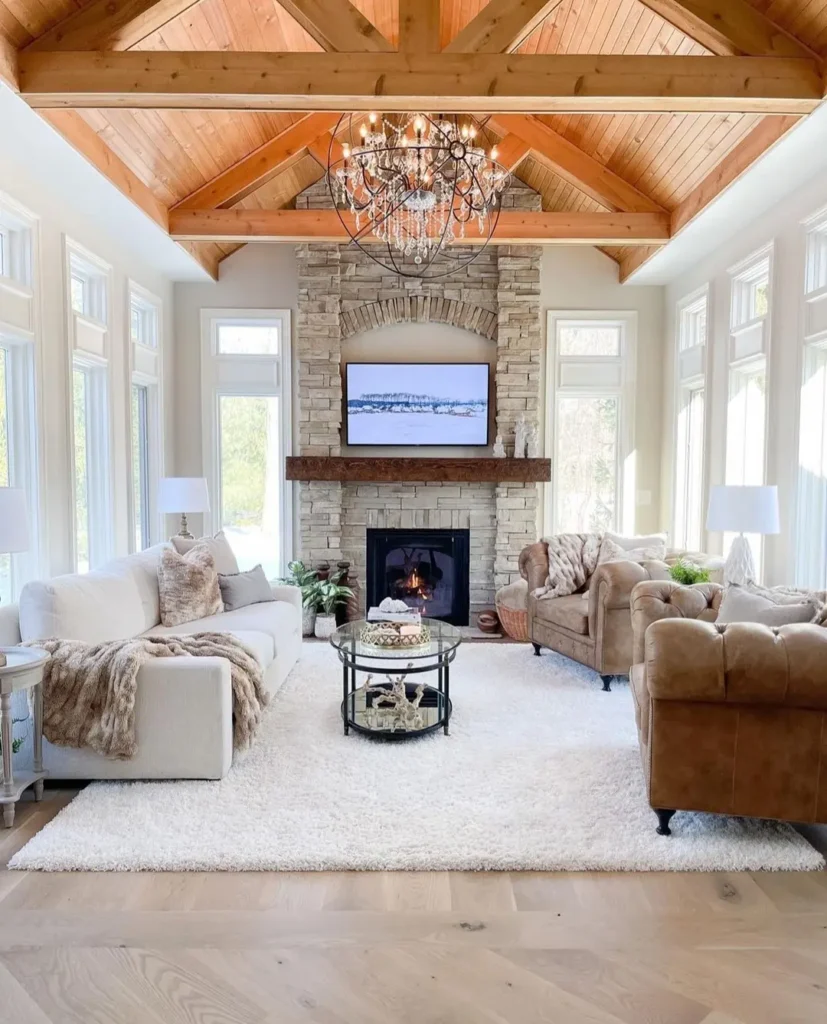 Bright rustic living room with vaulted wood ceiling, stone fireplace, neutral sofas, armchairs, a round coffee table, and large windows letting in natural light.
