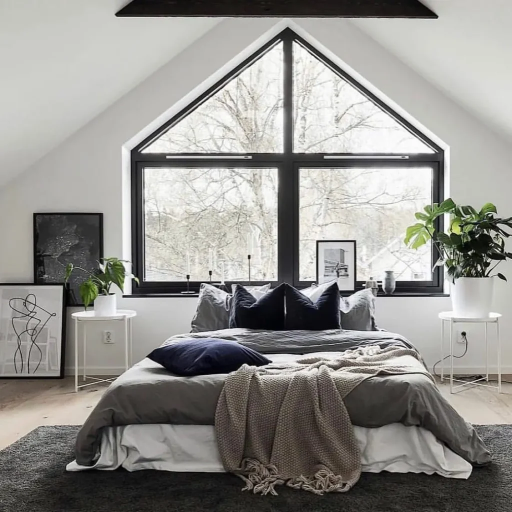 Scandinavian bedroom with a neutral color palette featuring a low-profile bed, layered gray and white bedding, black-framed triangular window, minimalist wall art, indoor plants, and a bright, airy modern bedroom design.