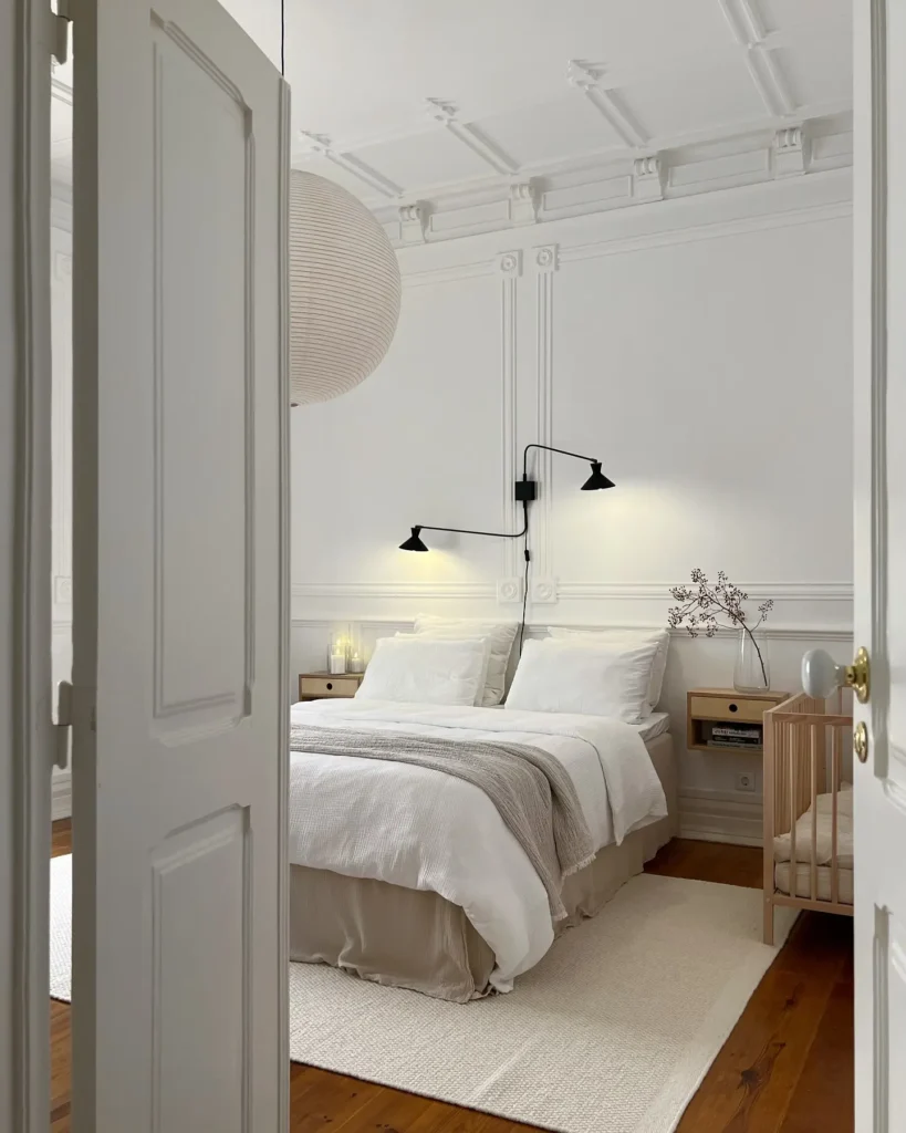 Minimalist Scandinavian bedroom featuring soft white walls, layered neutral bedding, wall-mounted black reading lights, natural wood nightstands, a light area rug, and an airy, calming modern bedroom design.