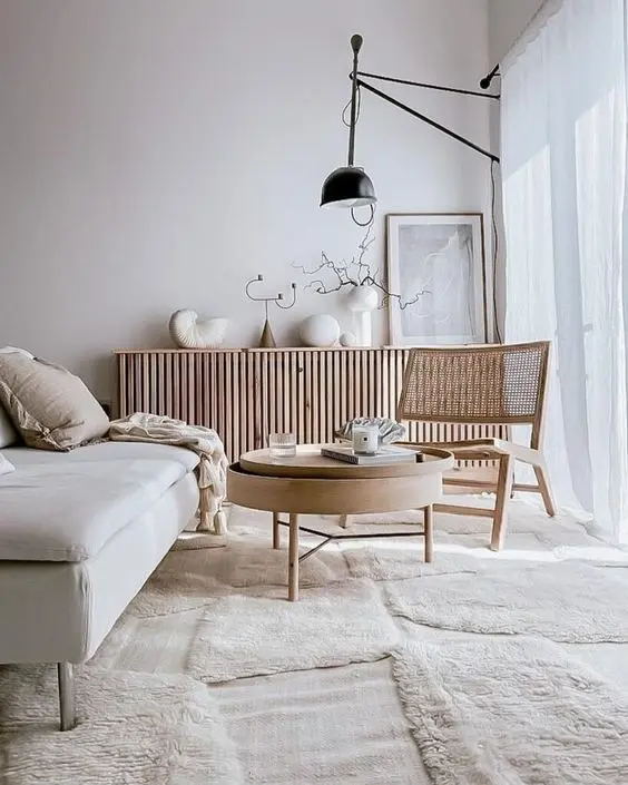 A minimalist Scandinavian living room with a white sofa, round wooden coffee table, cane chair, modern floor lamp, and a slatted wood sideboard against a white wall with decor items.
