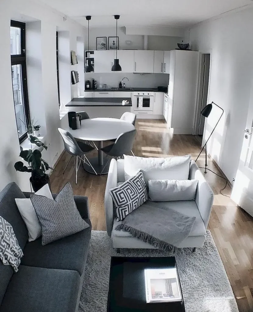 Modern open-plan studio apartment layout featuring a living room and kitchen with white walls, grey furniture, wooden floors, and minimalistic decor. A round dining table and pendant lights complete the space.