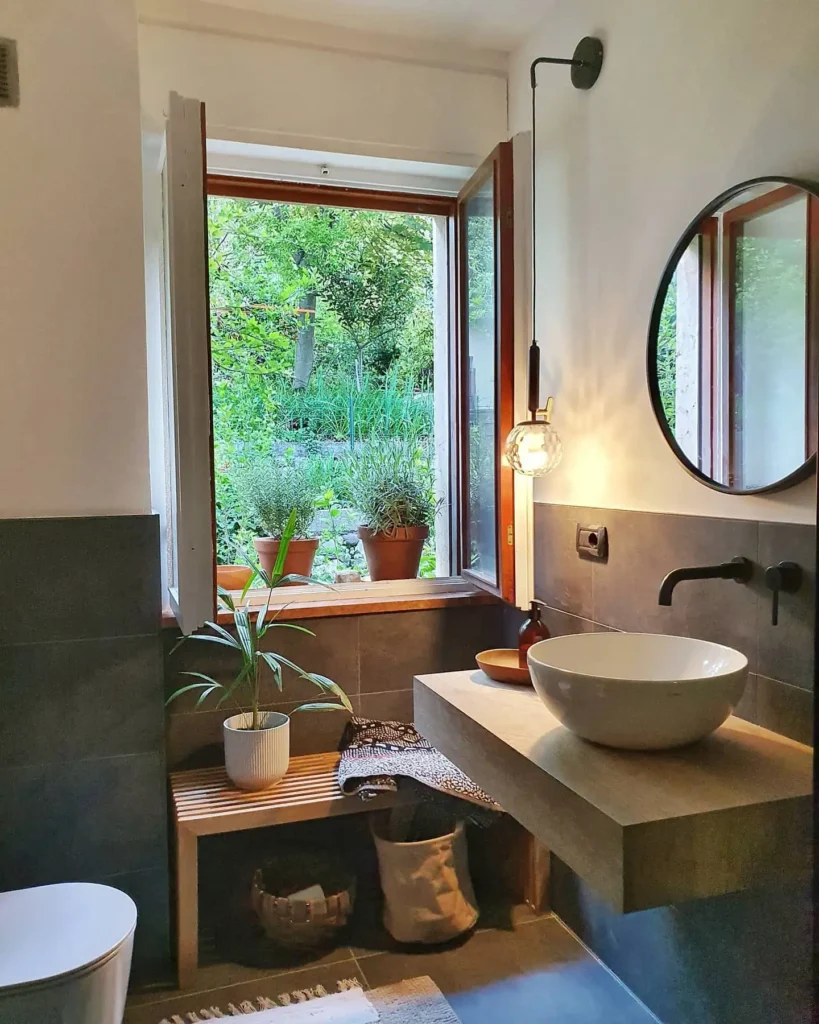 Small organic modern bathroom with a floating concrete vanity, round wall mirror, vessel sink, pendant light, indoor plants, and an open window bringing in natural light.