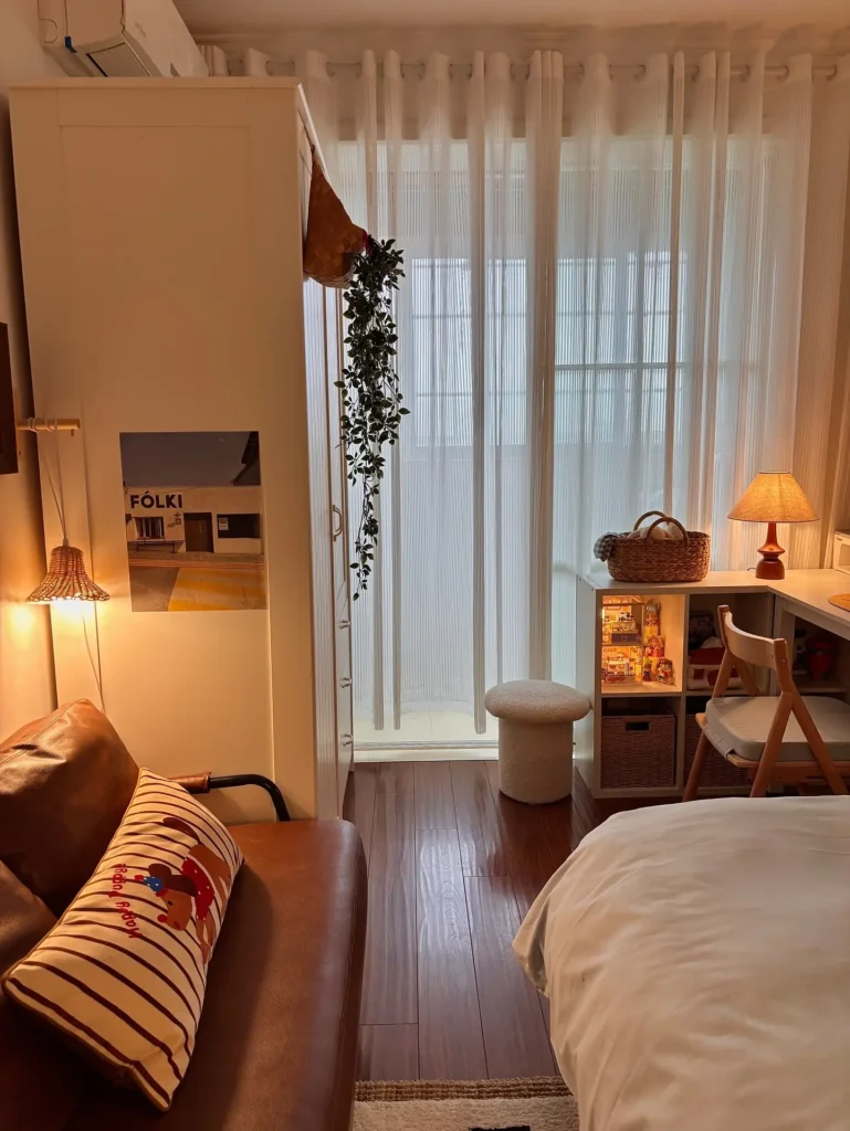 A cozy small room features a brown sofa, striped pillow, white wardrobe, sheer curtains, desk with lamp, and wooden flooring. Warm lighting creates an inviting atmosphere.
