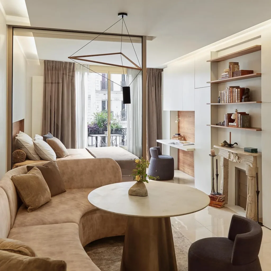 Modern studio apartment with a curved beige sofa, unifying color scheme, round table, bed, open shelves, faux fireplace, and a window with light curtains overlooking a small balcony with plants.
