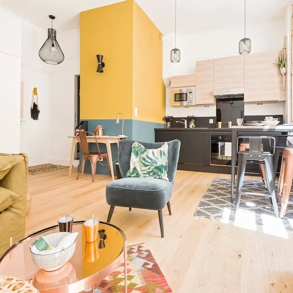 A modern studio open-plan living area features a gray armchair, yellow accent wall, black kitchen cabinets, wooden dining table, and colorful decor.