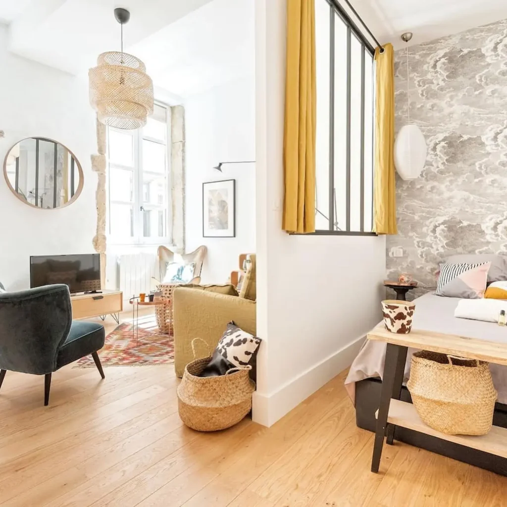 Bright, modern studio with a cozy living area, TV, bed, yellow curtains, and woven baskets on a light wood floor. Marble-patterned accent wall adds style to the bedroom space.