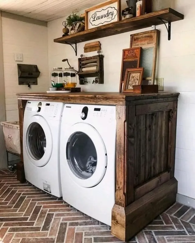 Two front-loading laundry machines are enclosed in a rustic wooden cabinet, with shelves above holding laundry supplies, vintage tools, and a "Laundry" sign for a charming farmhouse touch.