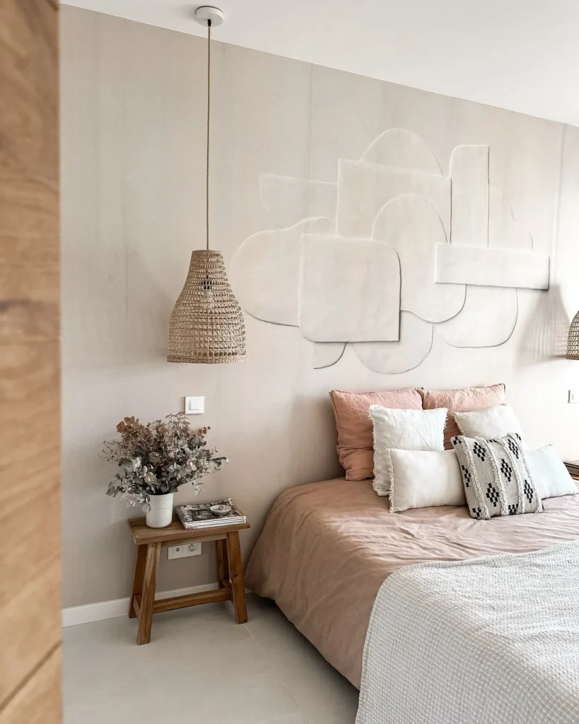 Wabi-sabi bedroom with a soft neutral accent wall, sculptural wall art, layered linen bedding, warm wood furniture, and a woven pendant light.