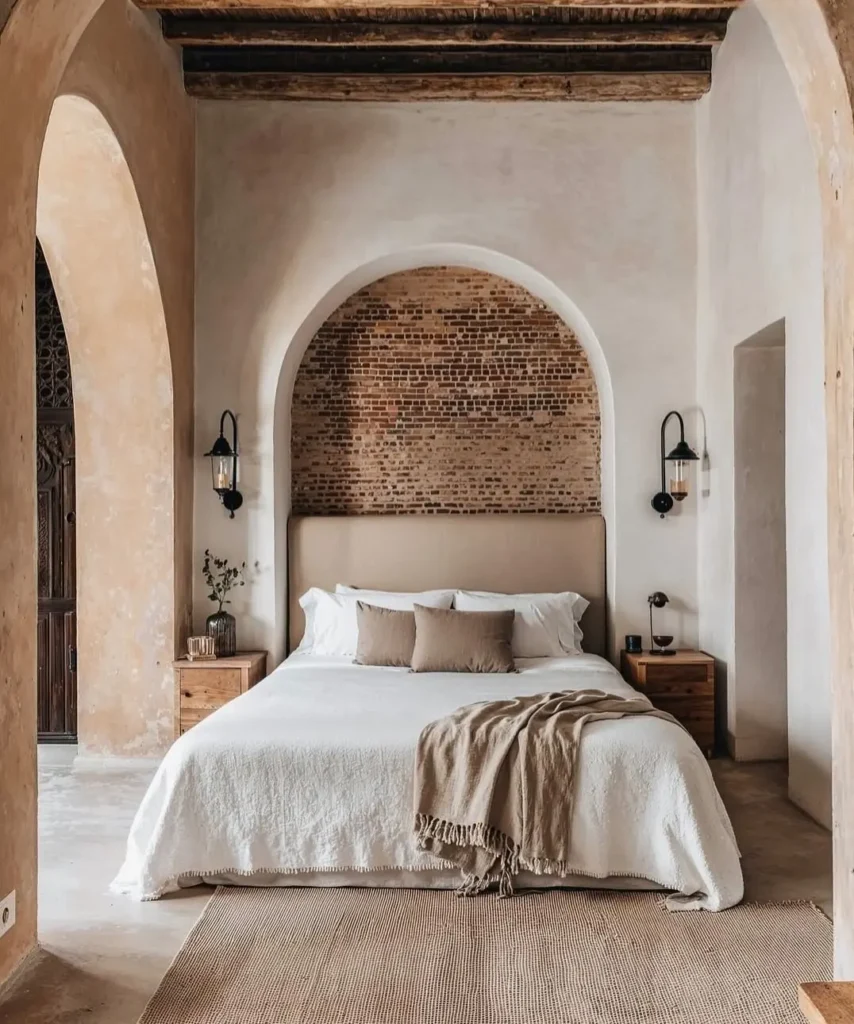 Wabi-sabi bedroom with arched plaster walls, exposed brick niche, natural wood ceiling beams, neutral bedding, and a warm, minimalist, earthy aesthetic.
