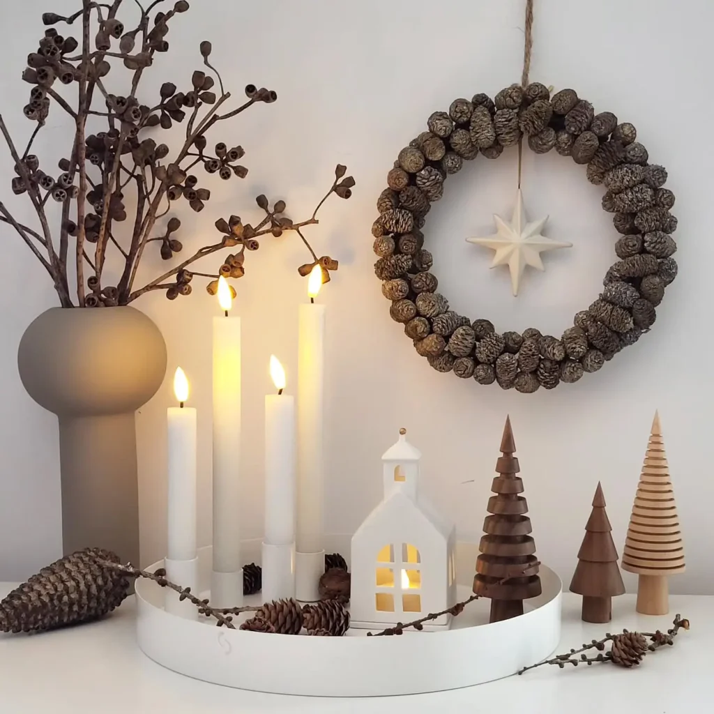 Minimalist winter holiday decor with lit candles, a ceramic house, wooden trees, a pinecone wreath, and a vase with branches on a crisp white surface.