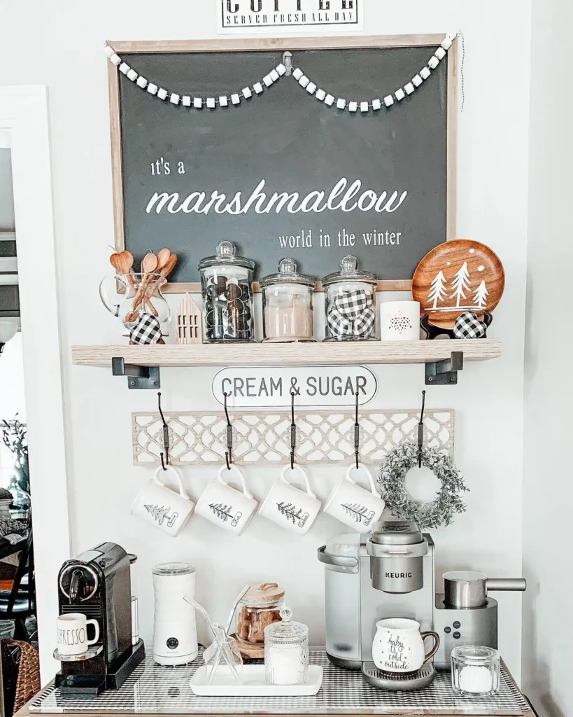 A winter coffee bar featuring mugs, coffee machines, a shelf with jars of treats, cozy winter-themed decor, and a sign reading "it's a marshmallow world in the winter.