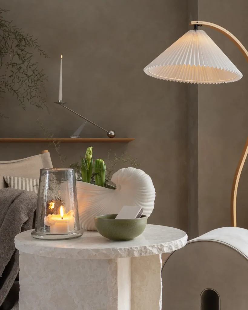 A round marble side table with a lit candle in a glass holder, a green bowl, white ceramic vase with flowers, and a pleated floor lamp creates cozy winter decor in a neutral-toned room.