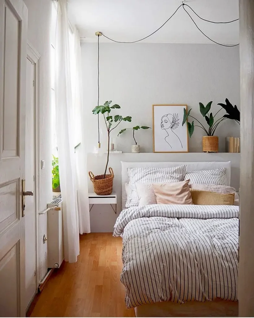 Small Scandinavian bedroom design featuring neutral striped duvet, potted plants, simple line art print, and light wood flooring.