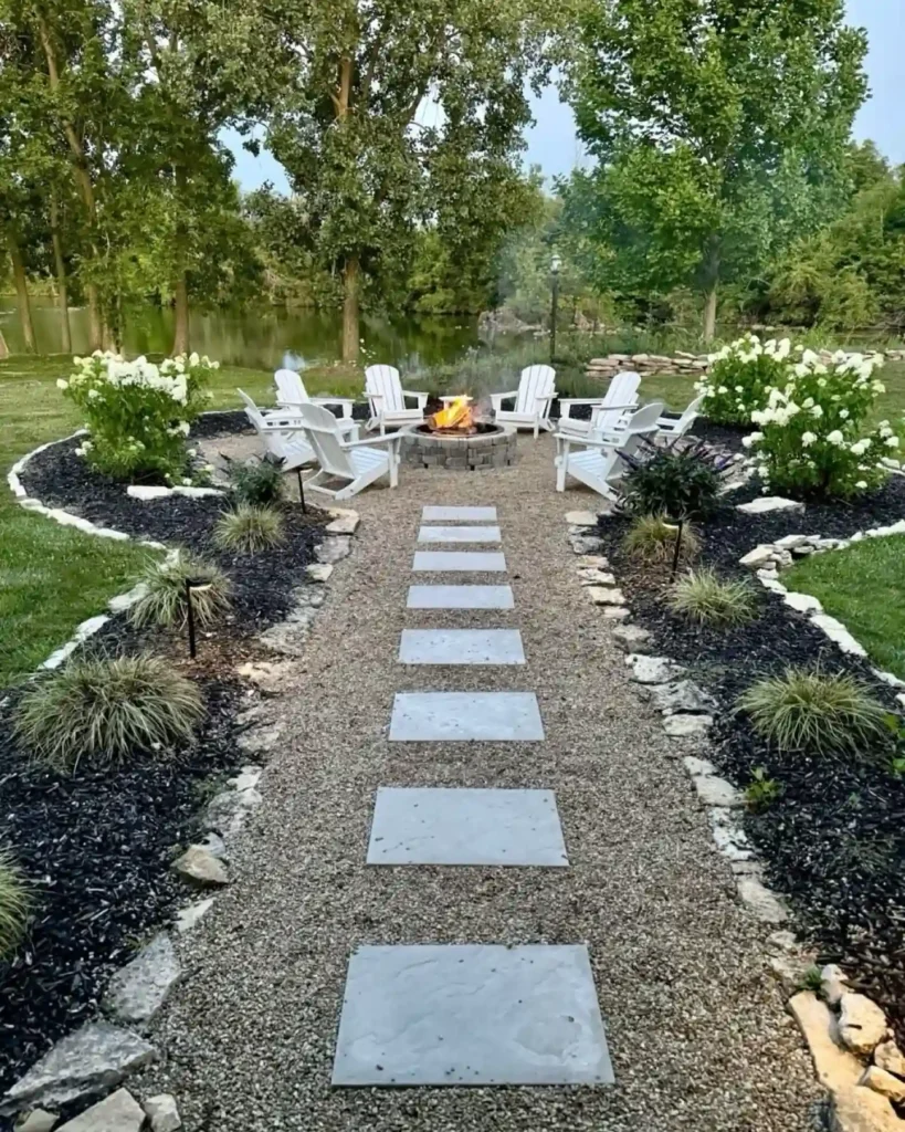 Gravel garden walkway with stone stepping stones leading to a circular fire pit patio with white Adirondack chairs and lush greenery.