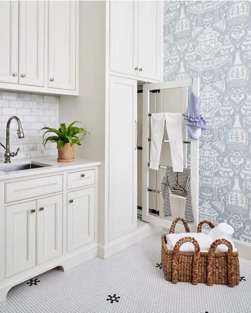 Small laundry room with built-in cabinet drying rack, white shaker cabinets, blue patterned wallpaper, woven laundry basket, and potted plant.