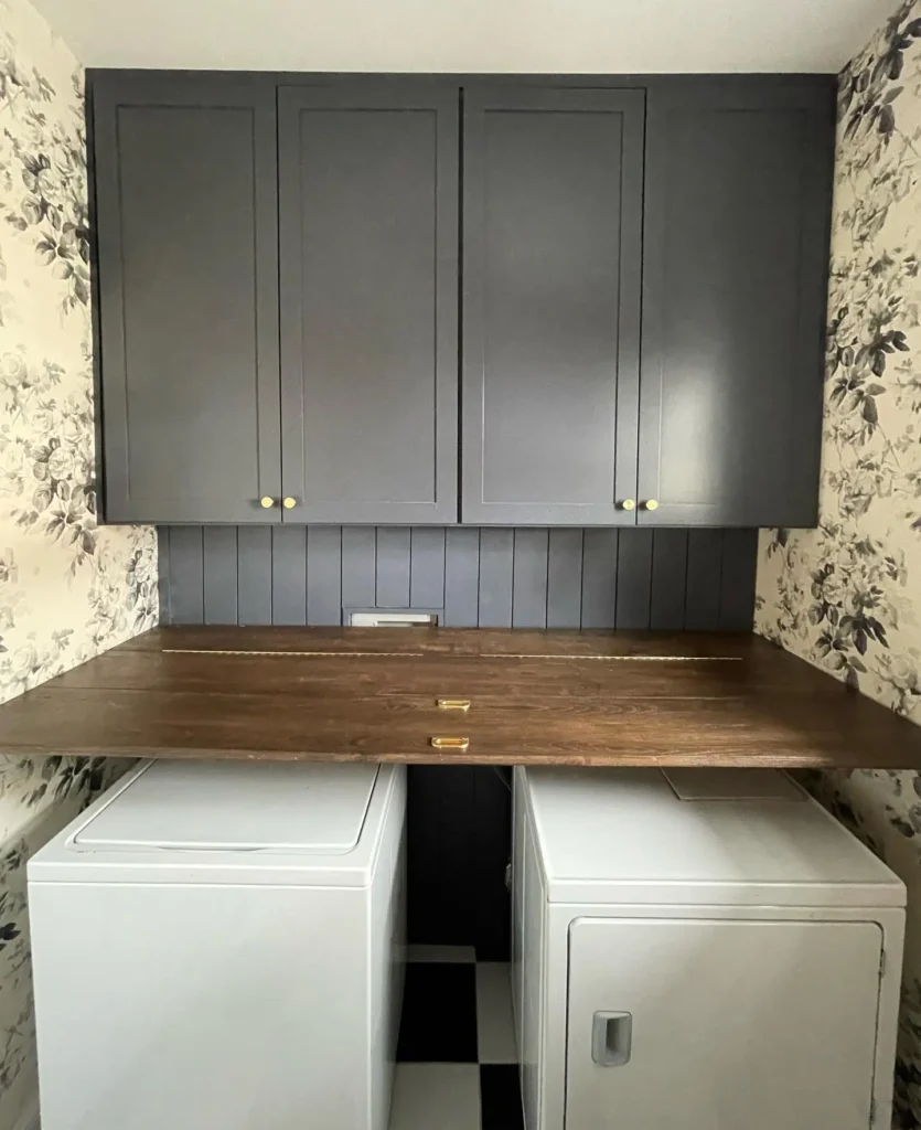 Laundry room with dark wood folding station and countertop, navy cabinets, floral wallpaper, and side-by-side washer and dryer.
