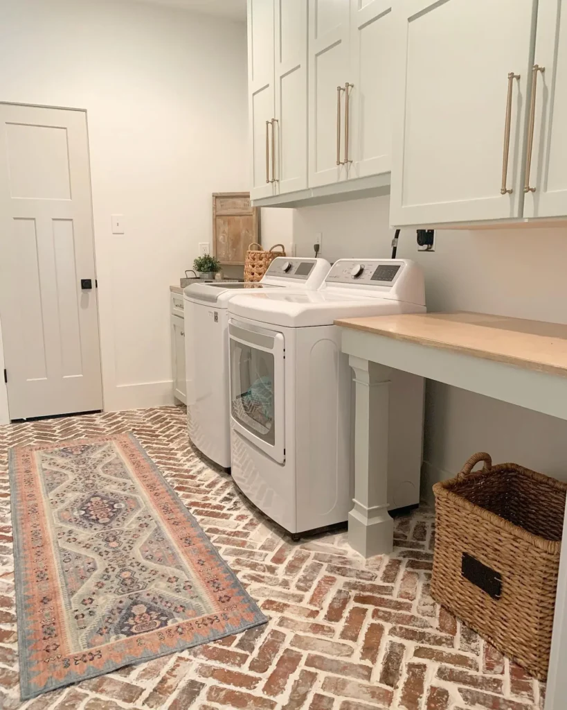 Modern farmhouse laundry room featuring brick herringbone flooring, white shaker cabinets, wood folding table, wicker basket, and patterned runner rug.