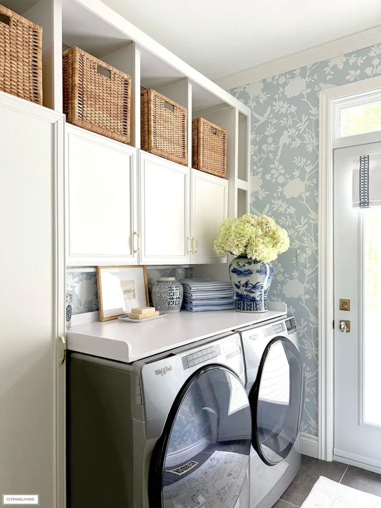 Small laundry room with white cabinets, wicker basket storage, blue floral wallpaper, front-load washer and dryer, and fresh hydrangeas on the countertop.