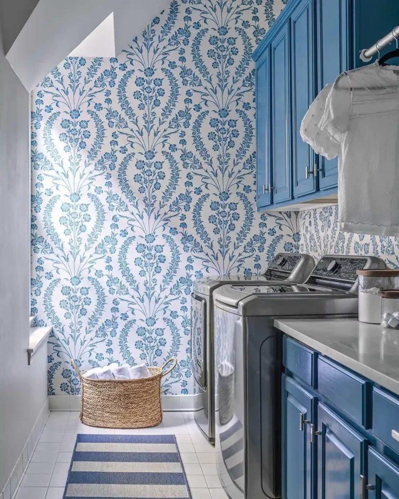 Blue floral wallpaper in small laundry room with navy cabinets, stainless steel washer and dryer, and woven laundry basket.