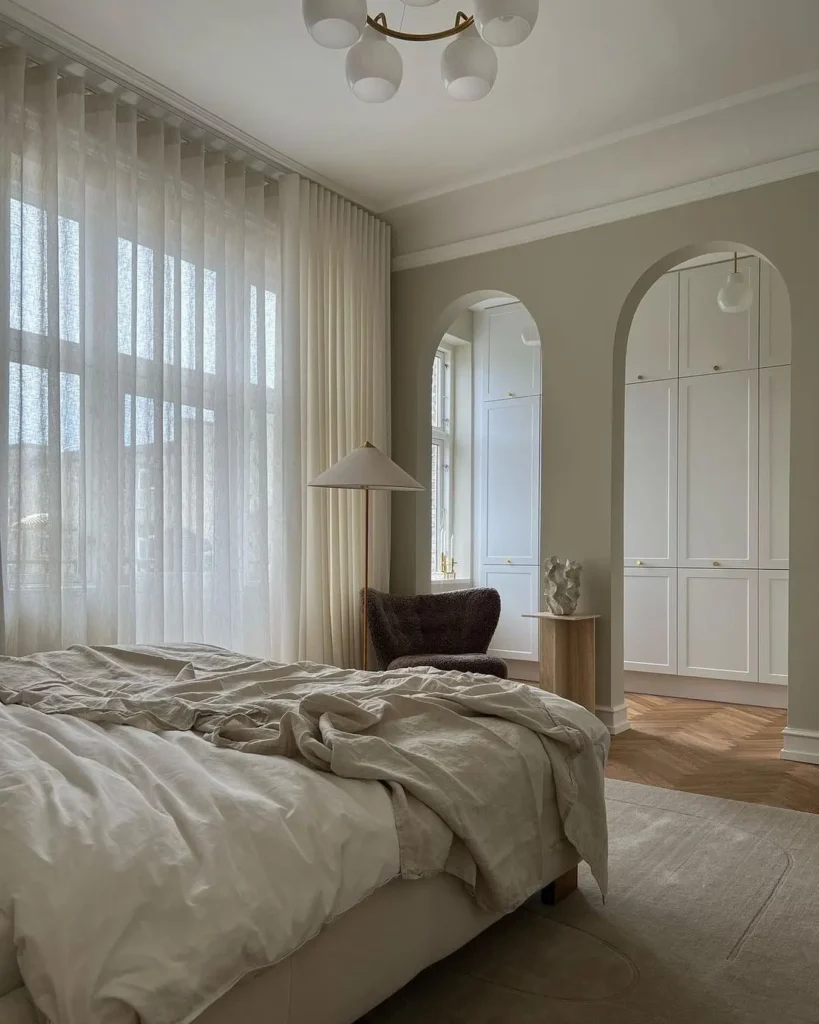 Minimalist bedroom with hidden storage, arched wall, neutral bedding, sheer curtains, and soft natural light.