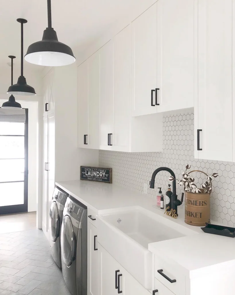 Modern white laundry room with shaker cabinets, farmhouse sink, hex tile backsplash, black hardware, and front-loading washer and dryer.