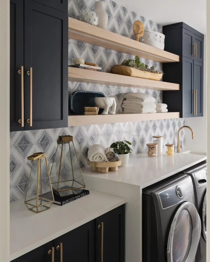 Modern laundry room storage with navy cabinets, light wood floating shelves, geometric tile backsplash, and front-loading washer and dryer.