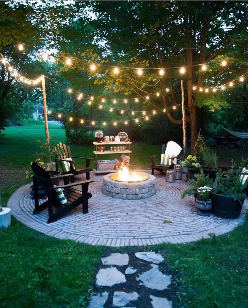Outdoor backyard seating area with stone fire pit, café string lights, wooden Adirondack chairs, and circular brick patio surrounded by trees.