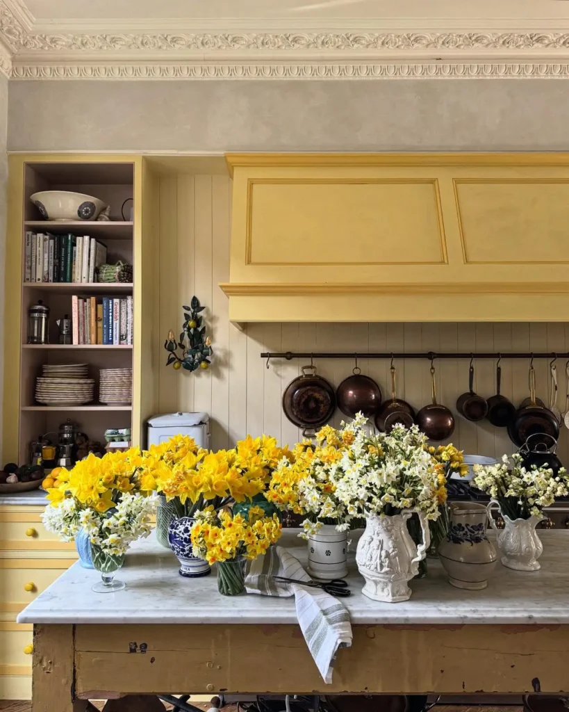 A kitchen with yellow cabinets, a marble-topped table filled with vases of yellow and white spring flowers, shelves with dishes, and copper pots hanging on the wall.