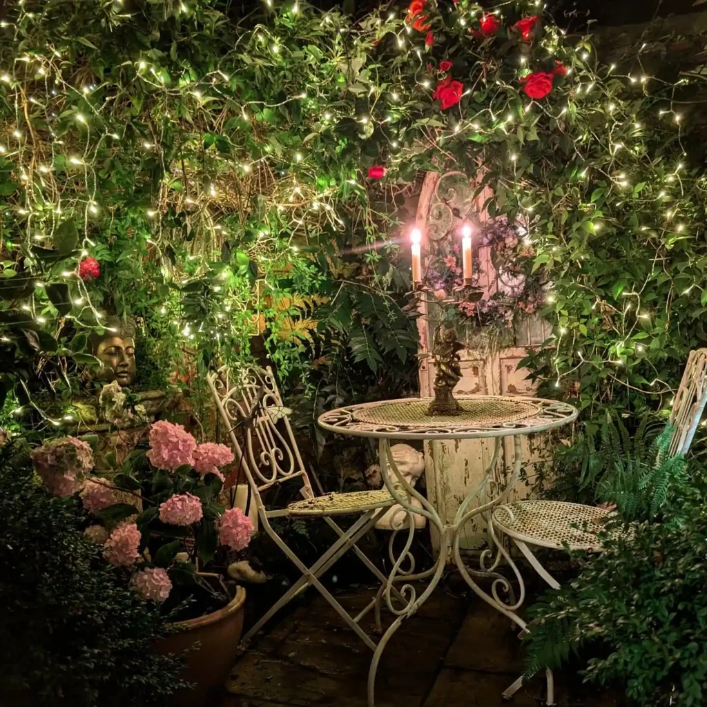 A small round table with two chairs is set in a lush spring garden, surrounded by greenery, pink flowers, string lights, and two lit candles on the table.