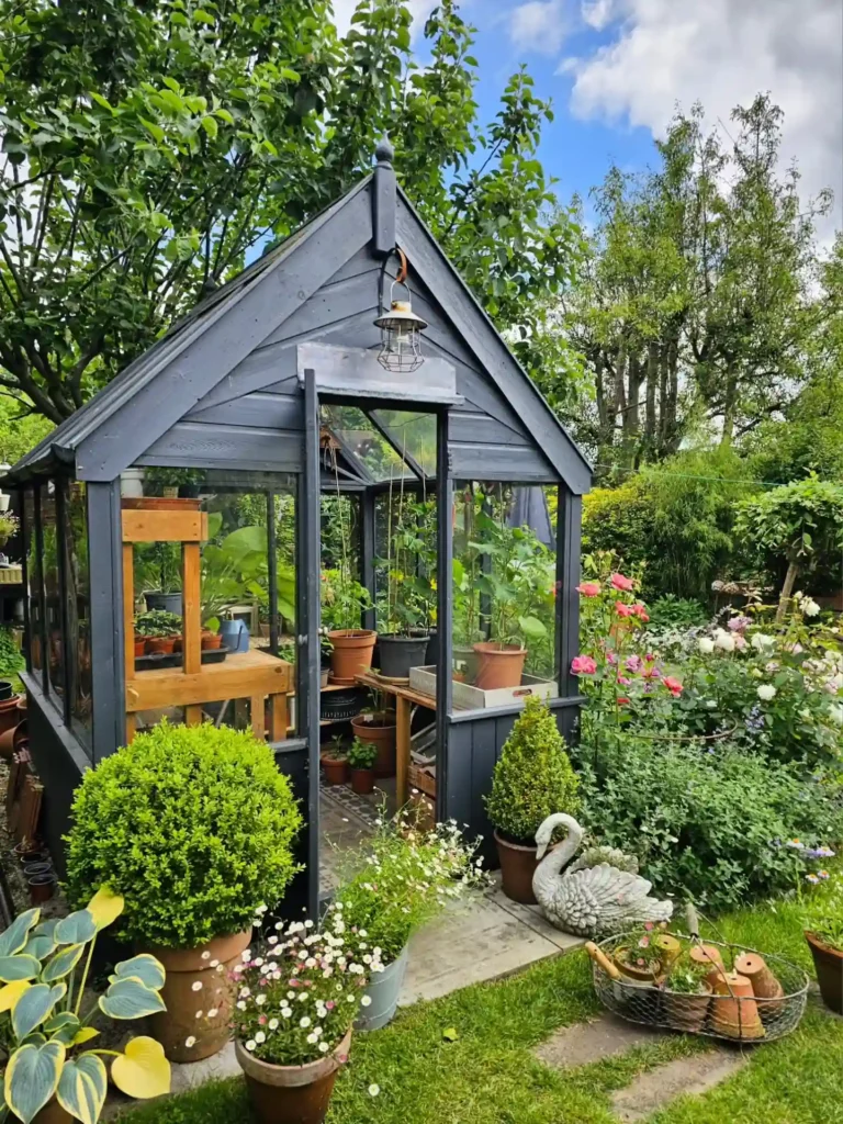 A small glass greenhouse with potted plants inside and around it, nestled in a lush spring garden filled with flowers, bushes, and a decorative swan statue.