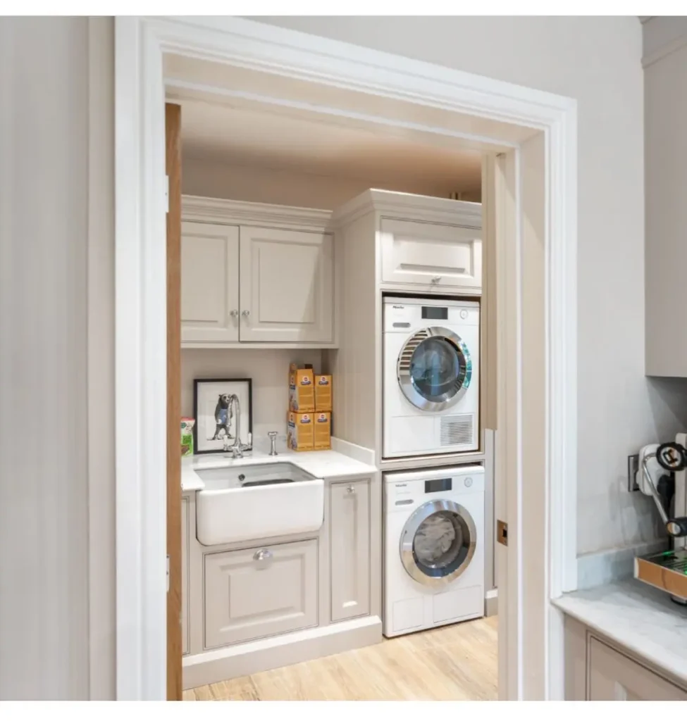Small laundry closet featuring stacked washer and dryer, apron-front sink, upper cabinetry, and neutral cabinetry in a narrow space.