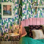 A stylish bedroom featuring a multicolored geometric wallpaper, pink tufted headboard, and decorative green bedding, with a framed poster and shelves on the wall.