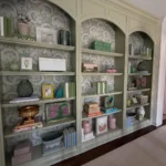 Floor-to-ceiling sage green built-in bookshelves with patterned wallpaper backing, styled with various decorative objects, books, and framed art.
