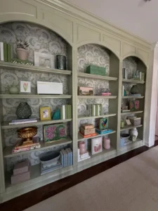 Floor-to-ceiling sage green built-in bookshelves with patterned wallpaper backing, styled with various decorative objects, books, and framed art.