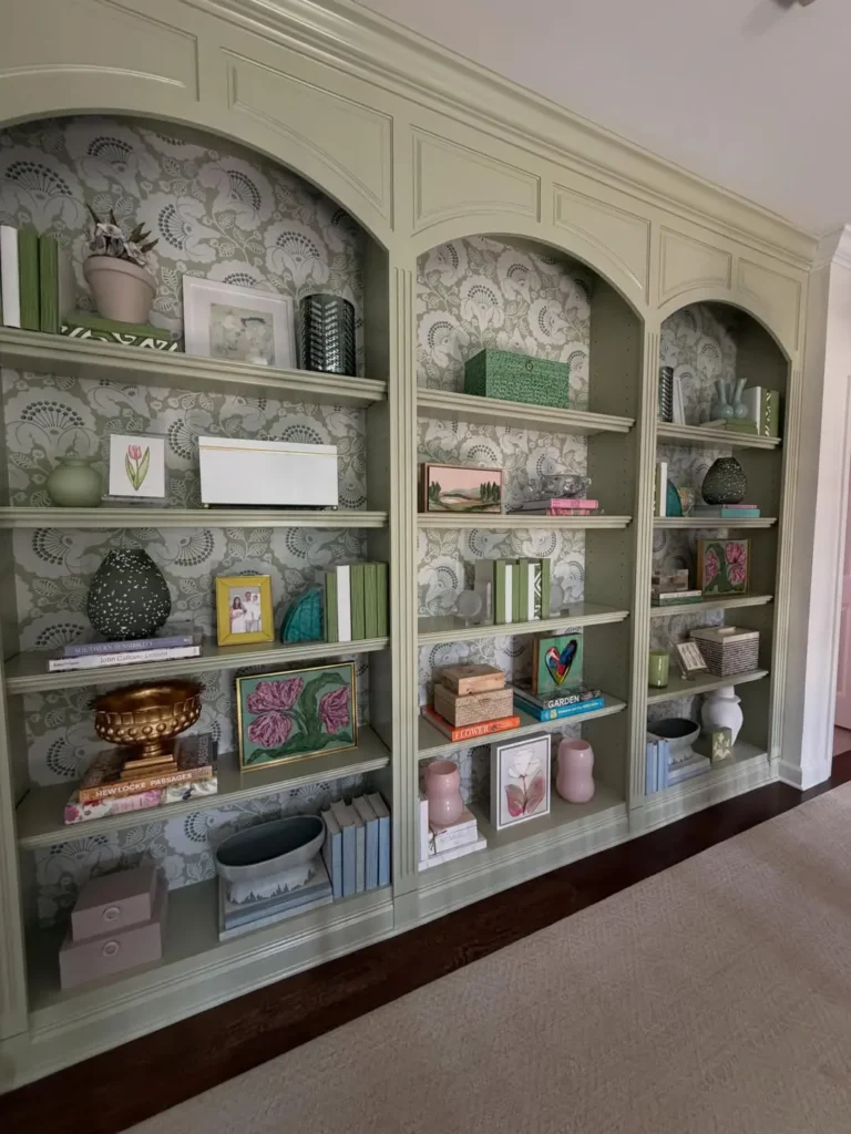 Floor-to-ceiling sage green built-in bookshelves with patterned wallpaper backing, styled with various decorative objects, books, and framed art.